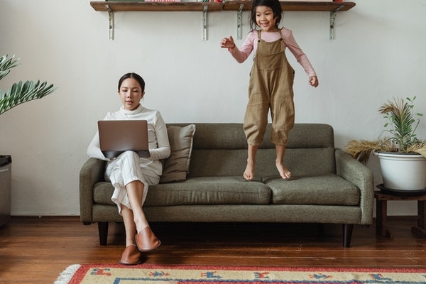 Child jumping on a couch while parent works off a laptop.