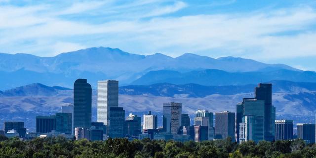 Steel gray skyscrapers stand in the foreground of a deep blue mountain range in Denver, host city of the Colorado Mortgage Expo