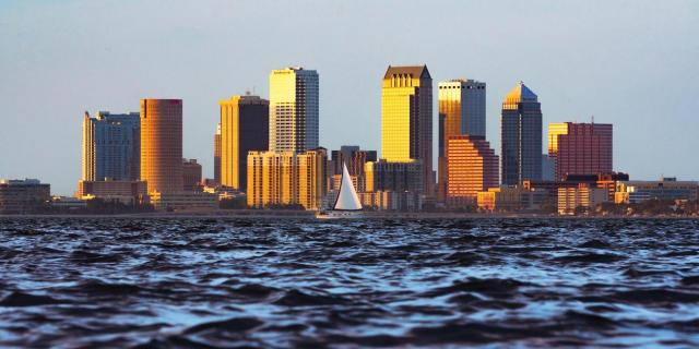 A view of Tampa, host of the Suncoast Mortgage Expo, as seen from the ocean shining in the setting sun, with a single white sailboat standing in front of the city skyline.