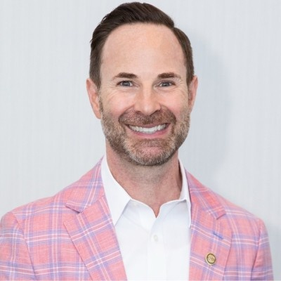 White man with brown hair, a short beard, a white collared shit and a pink and blue plaid sport coat.