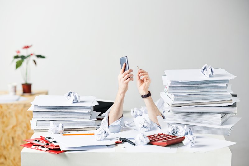 A loan officers arm reach up from beneath a pile of papers and work.